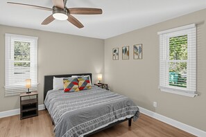 A bedroom with a bed, a ceiling fan, two bedside tables with lamps, and three framed botanical prints on the wall.
