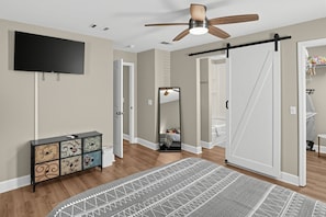 Quarto com ventoinha no teto, TV de tela plana, penteadeira com gavetas decorativas, tapete estampado e porta de guarda-roupa deslizante.
