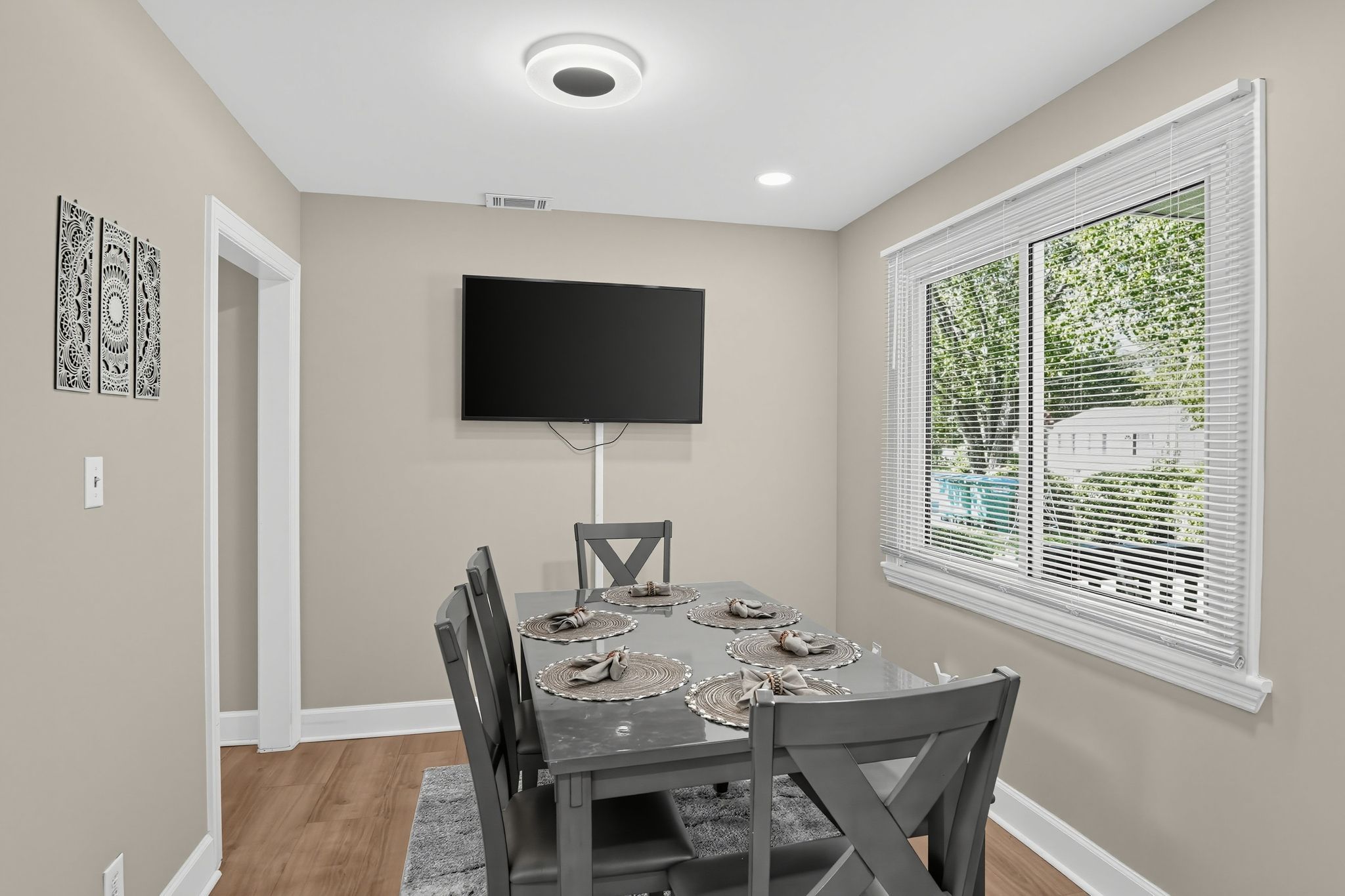 A dining area with a table set for four, a flat-screen TV mounted on the wall, and a window with blinds.