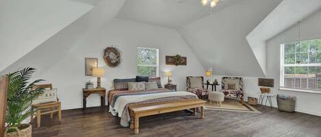 A spacious attic bedroom with a large bed, a wooden bench, a ceiling fan, and a view of the outdoors through a window.