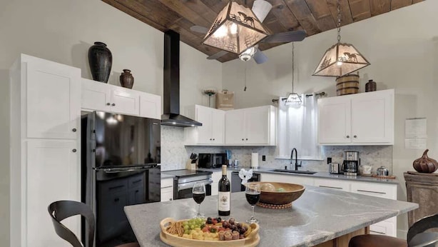 A modern kitchen with white cabinets, a stainless steel refrigerator, a wooden island with a fruit platter and wine glasses, and pendant lighting. - Casa Blanca-swim Spa-steps to Main Street! (Fredericksburg)