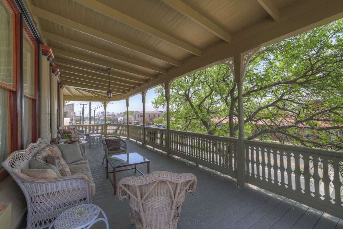 A covered porch with wicker furniture, a railing, and a view of trees and buildings.