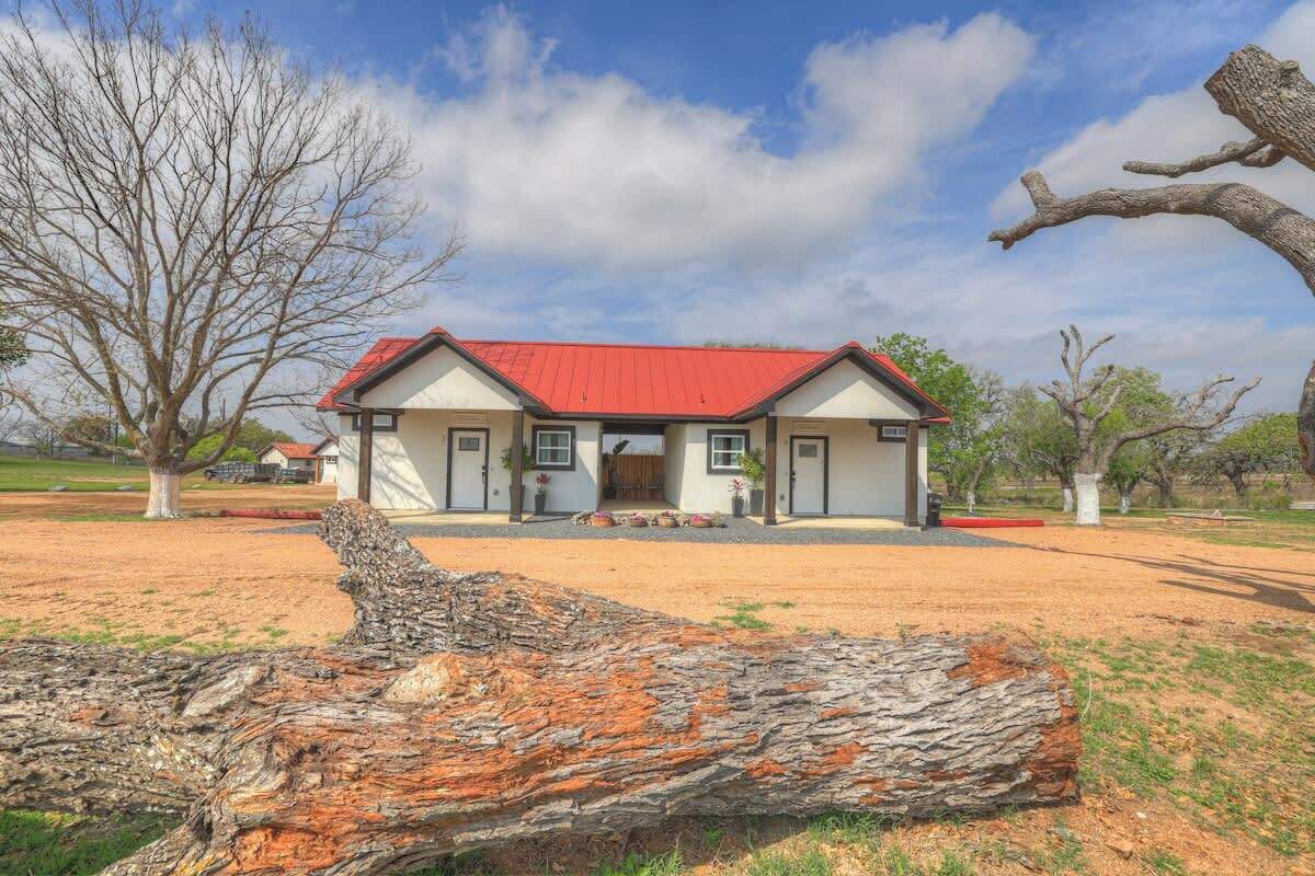 Une maison avec un toit rouge, un porche à l’avant et un grand arbre au premier plan.
