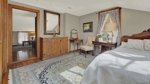 A bedroom with a large bed, a wooden dresser, a chair, a mirror, and a window with curtains.