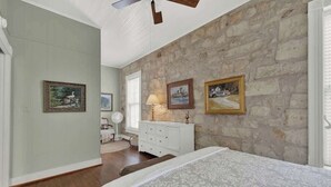 A bedroom with a stone wall, a bed, a dresser, and framed pictures. - Fountain Suite-stay Right on Historic Main Street in the Heart of Fredericksburg (Fredericksburg)