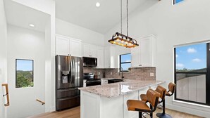 A modern kitchen with white cabinets, a large island with bar stools, stainless steel appliances, and a hanging light fixture. - Hill Country Escape W/views Plus, Hot-tub and Pet Friendly! (Kerrville)