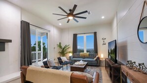 A modern living room with a ceiling fan, large windows, a sofa, a coffee table, and a TV.