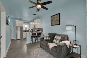 A modern kitchen with a central island, a dining area with bar stools, a grey sofa with patterned cushions, and a ceiling fan.