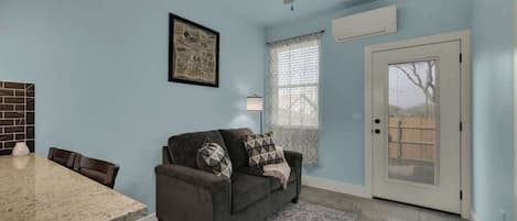 A room with a dining table, a sofa, a ceiling fan, a window with blinds, and a door.