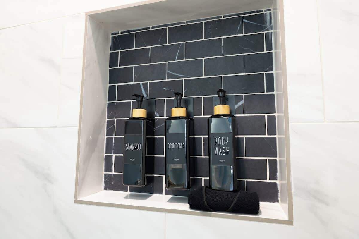 A bathroom with a black and white tiled wall, three bottles of body wash, shampoo, and conditioner, and a black towel.