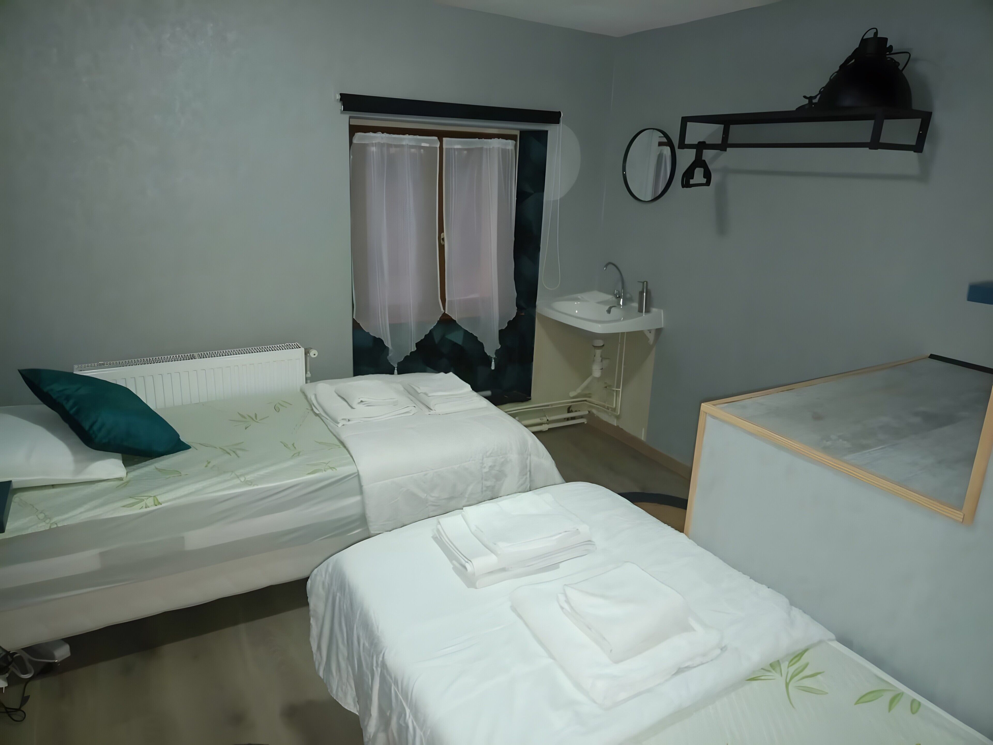 Ein Schlafzimmer mit zwei Betten, einem Fenster mit Vorhängen, einem Nachttisch, einem Wandregal und einem kleinen Waschbecken.