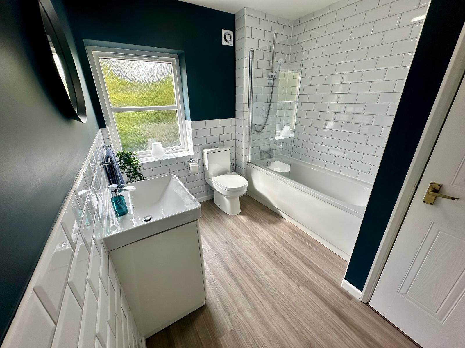 A modern bathroom with a white sink, toilet, and bathtub, featuring a window with a view of greenery.