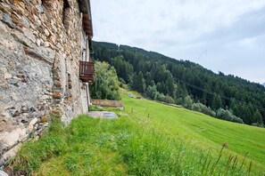 Un bâtiment en pierre doté d’un balcon en bois, un champ verdoyant et, au loin, une colline boisée.