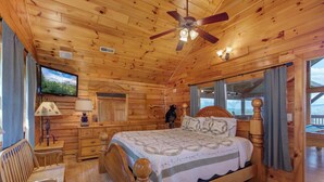 Ein Schlafzimmer in einer Holzhütte mit Bett, Stuhl, Schreibtisch, Deckenventilator und Blick ins Freie.