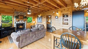 Un intérieur de chalet chaleureux avec une cheminée en pierre, des poutres en bois au plafond, un canapé, une table à manger et un téléviseur.