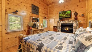 Une chambre dans un chalet en rondins, avec une cheminée en pierre, un lit recouvert d’une courtepointe à motifs et un téléviseur fixé au-dessus de la cheminée.
