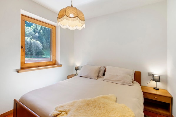 A bedroom with a bed, a window with a view of trees, a bedside lamp, and a hanging light fixture.