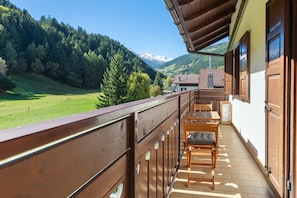 A balcony with a wooden railing, a table with chairs, and a view of a green valley and mountains.