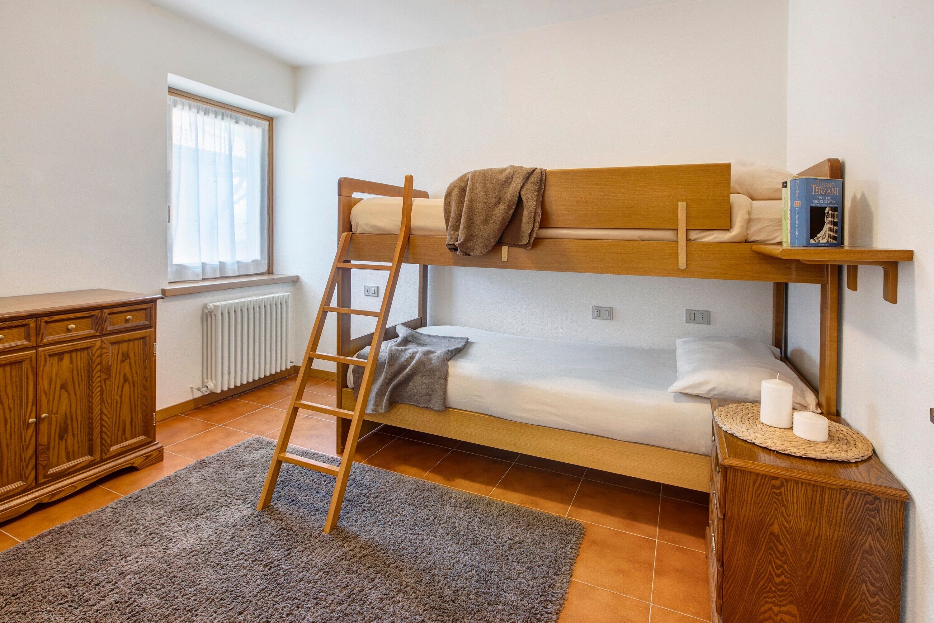 A bunk bed room with a ladder, a bedside table with candles, a book, and a window with curtains.