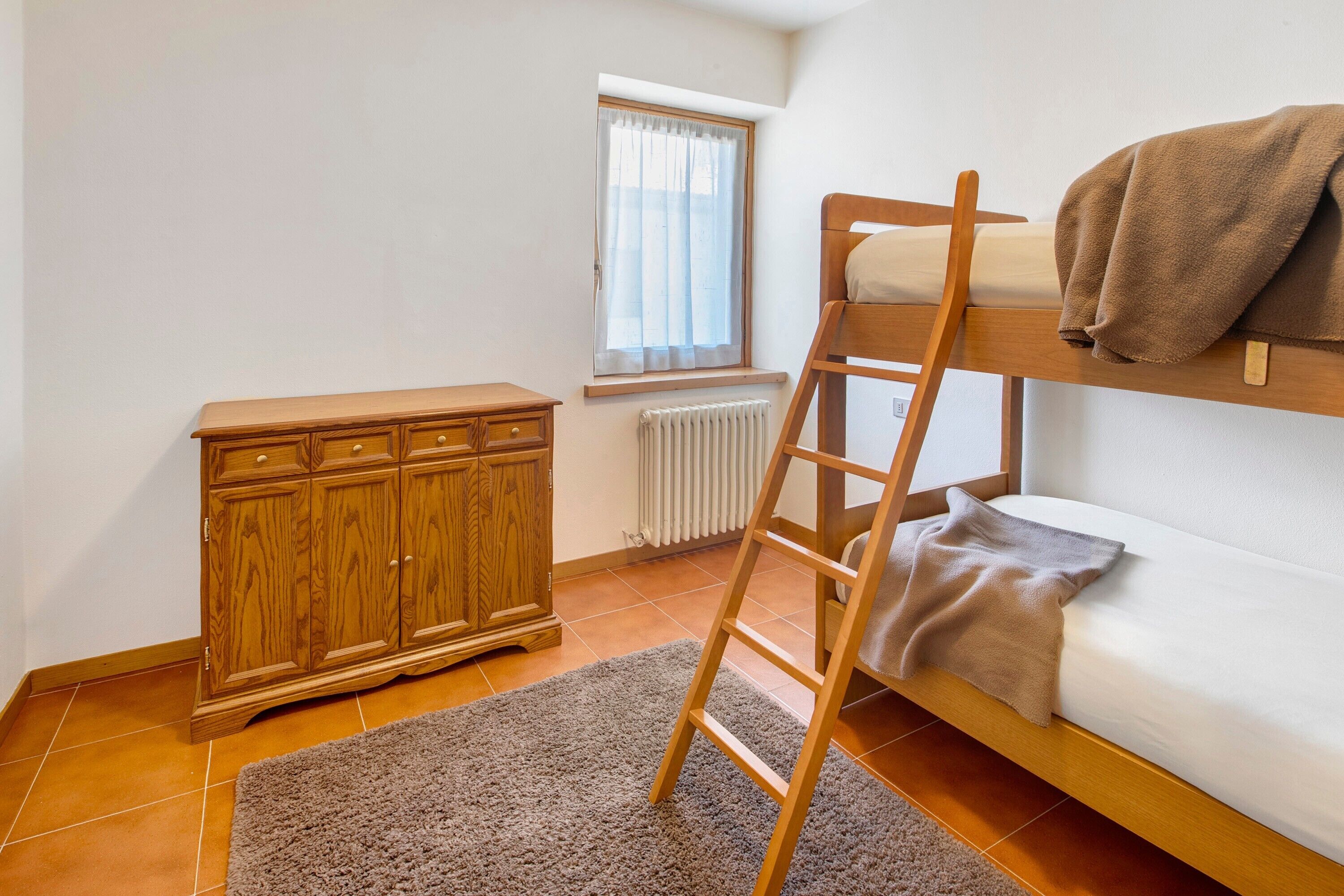 A room with a bunk bed, a wooden cabinet, a radiator, and a window with curtains.