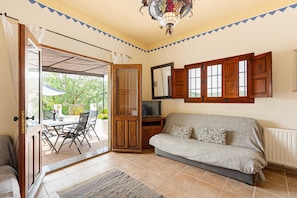 An open door leading to an outdoor area with a table and chairs, a sofa inside, a television on a stand, and a decorative ceiling light.