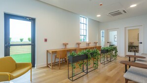 A modern interior with wooden desks, chairs, and a yellow armchair. There are potted plants on the desks and a large window with a view of greenery outside. - Studio 613 at The Frank (Birmingham)