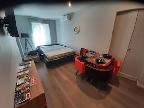 Une pièce de taille modeste comprenant un lit, une table à manger avec des chaises, un petit bureau avec des livres et une fenêtre avec des rideaux.