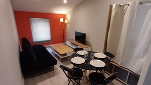 Un espace de vie compact comprenant un coin repas, un canapé, un téléviseur et une fenêtre avec des stores.