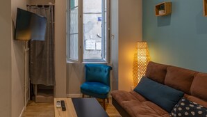 A room with a blue chair, a brown sofa, a TV mounted on the wall, a window with a view, and a lamp. - Appart 100 - Cosy T2 with TV (Bourg-Saint-Andéol)