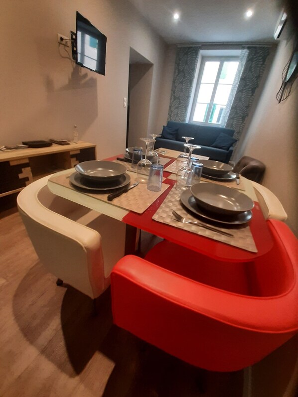 Un coin repas avec une table dressée pour quatre personnes, des chaises, un téléviseur fixé au mur et une fenêtre avec des rideaux.