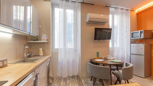 A compact kitchen with a sink, microwave, and refrigerator, a dining table with chairs, and a TV mounted on the wall.