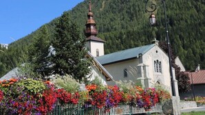 A church with a bell tower, surrounded by a fence with colorful flowers, set against a forested hillside.