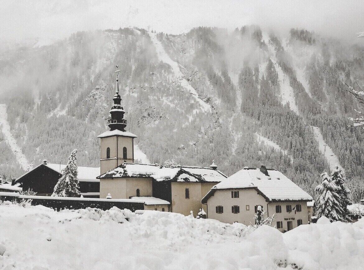 Un poble cobert de neu amb una església i edificis, envoltat de muntanyes.