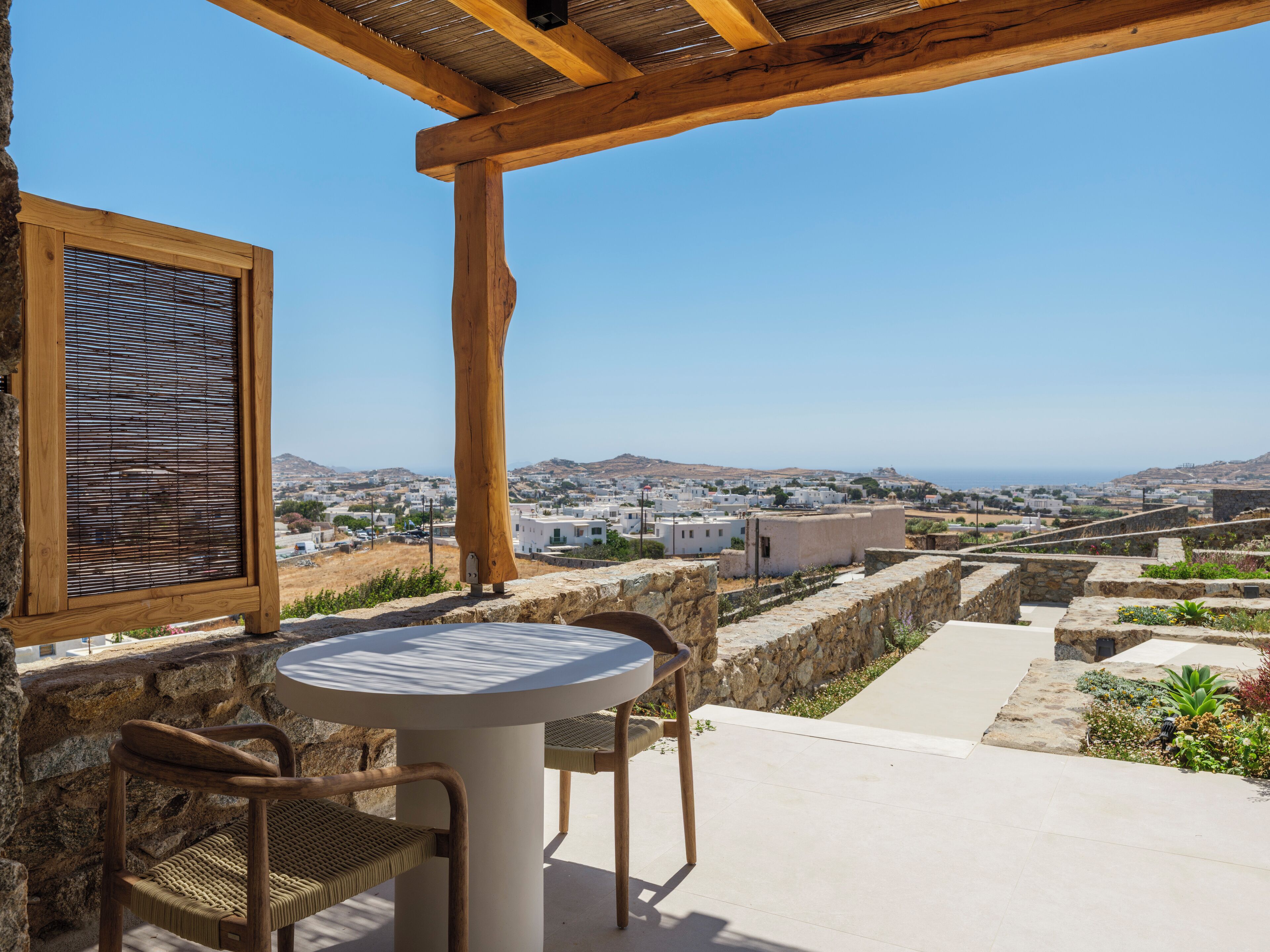 A patio with a round table and two chairs, overlooking a coastal town with white buildings and a clear blue sky.