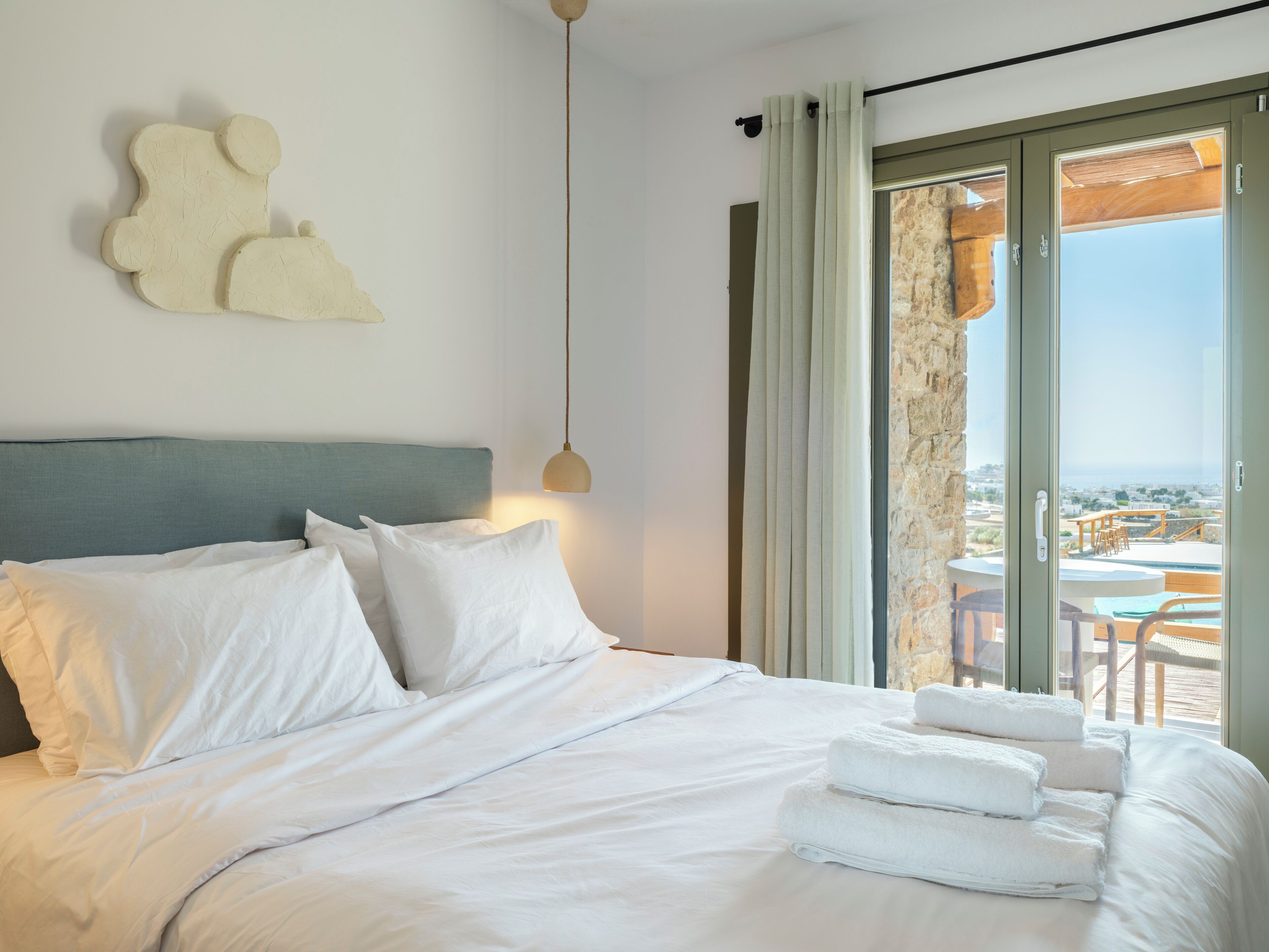 A neatly made bed with white linens, a grey headboard, and a view of the outdoors through a sliding glass door.