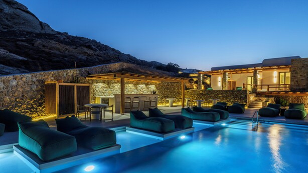 A poolside area with outdoor seating and a bar, surrounded by stone walls and a mountain backdrop. - Mykonian Shades (Ano Mera)