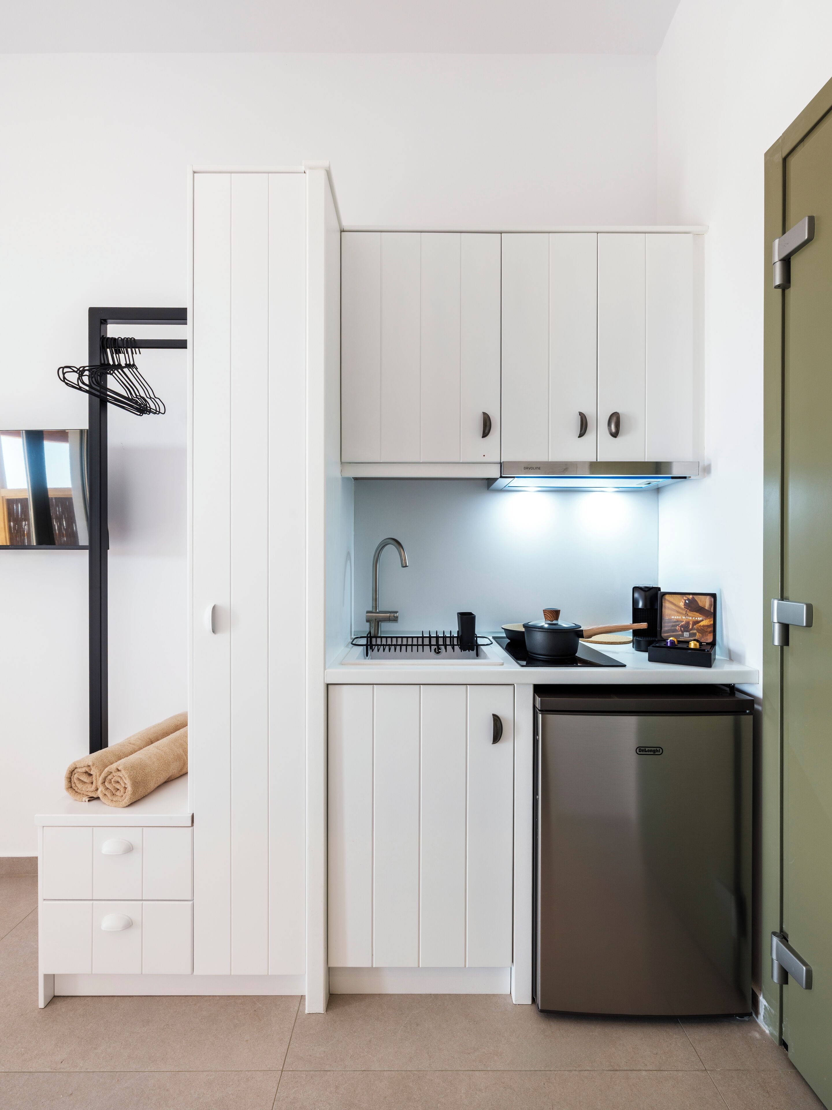 A compact kitchen with white cabinets, a stainless steel refrigerator, a sink, and a small countertop with a kettle and a coffee machine.