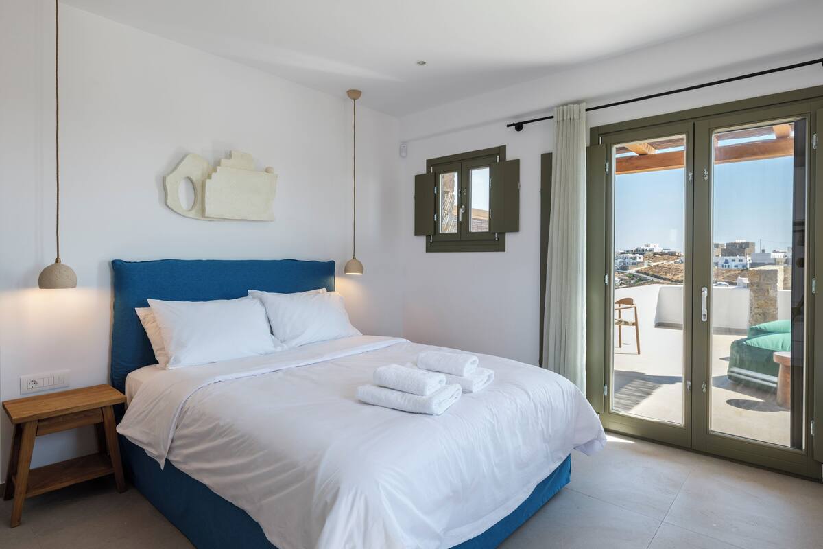 Un dormitorio con cama, mesitas de noche, vistas a través de un ventanal corredero y una ventana con contraventanas.