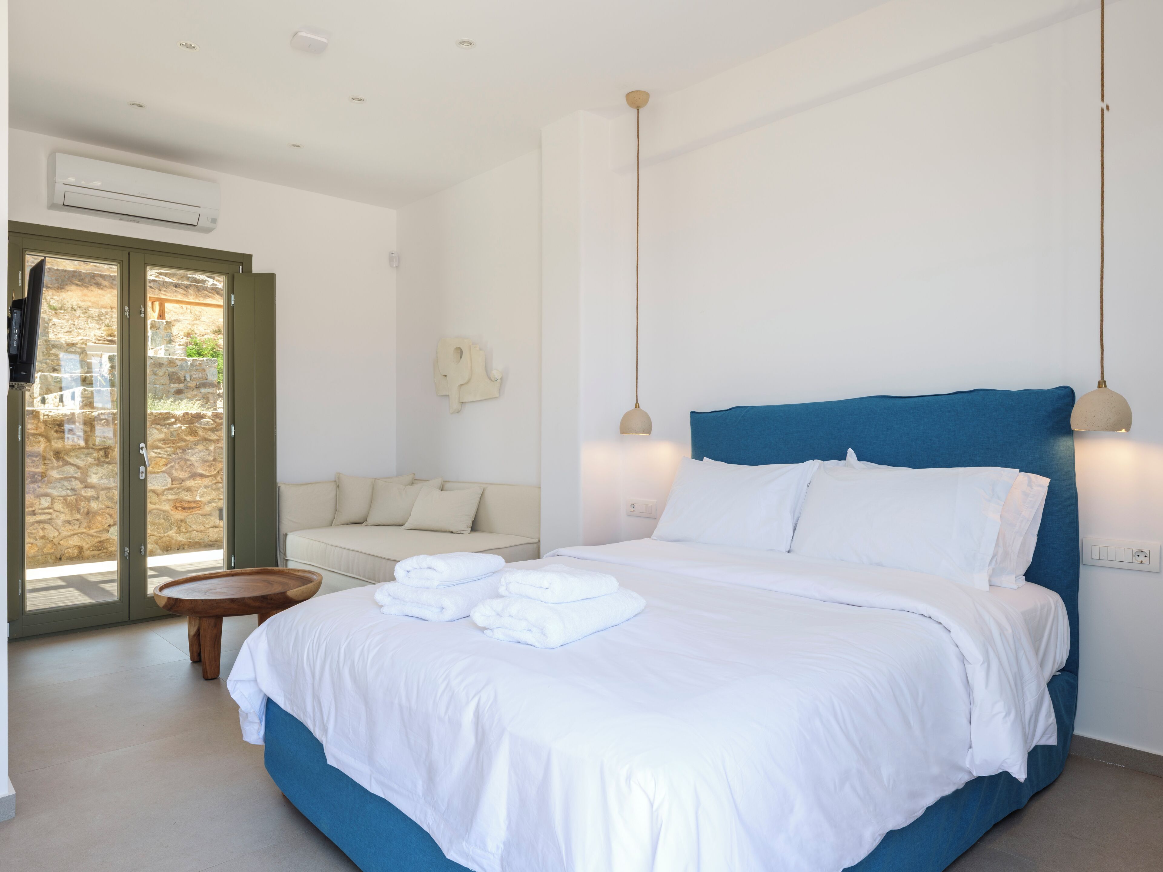 Un dormitorio con cama, sofá y una mesita. Se ve una ventana con vistas a un muro de piedra y follaje.