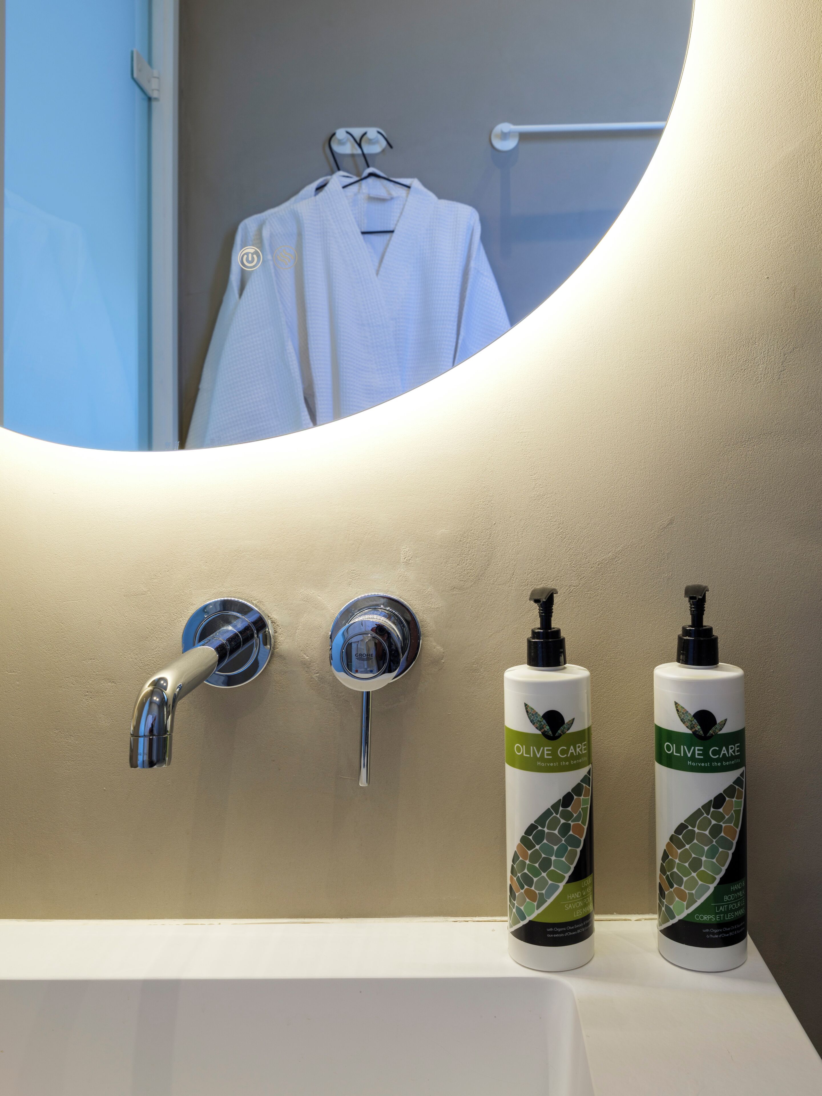 A bathroom with a white sink, two olive care hand wash bottles, a chrome faucet, and a mirror reflecting a white robe on a towel rack.