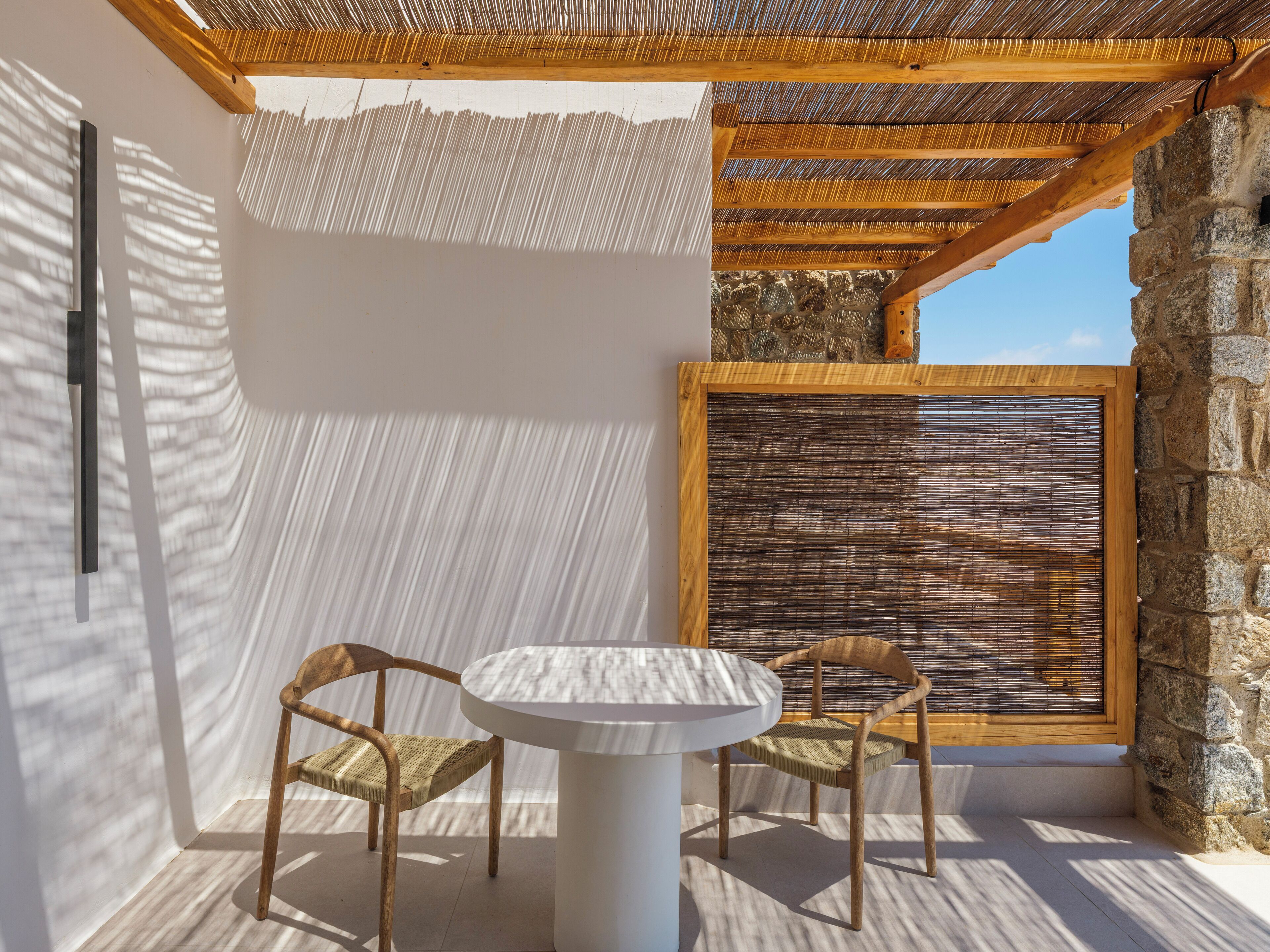 A sunlit patio with a round table and two wicker chairs, a stone wall, and a wooden ceiling structure.