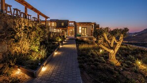 A modern house with a paved pathway leading to the entrance, surrounded by landscaped gardens and a view of the countryside. - Mykonian Shades (Ano Mera)