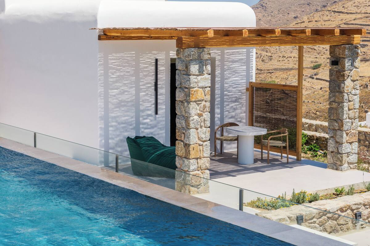 Un área junto a la piscina con un muro de piedra, una pérgola de madera y una mesa con sillas.