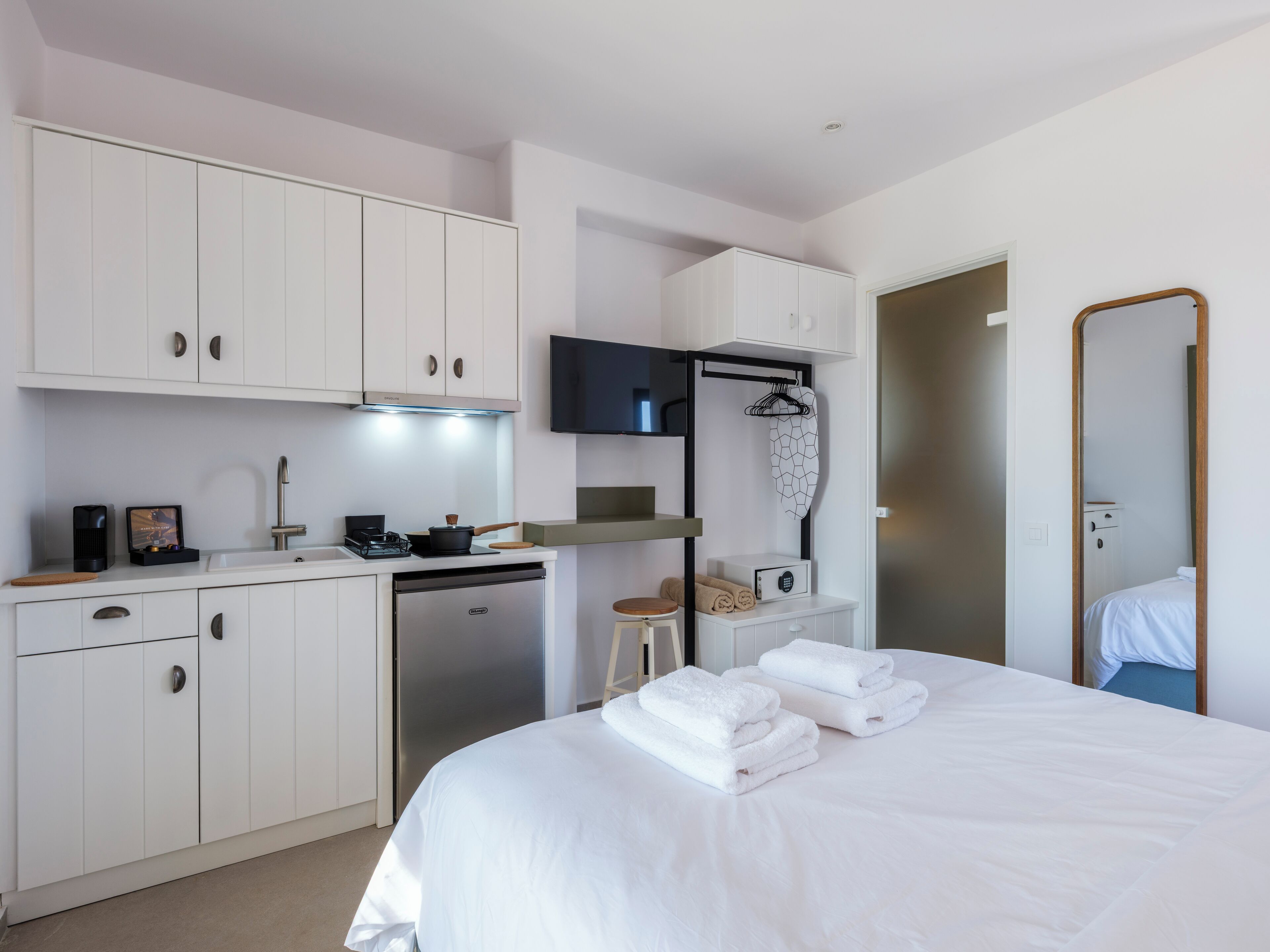 A modern hotel room with a kitchenette, a bed with neatly folded towels, a TV, and a mirror.