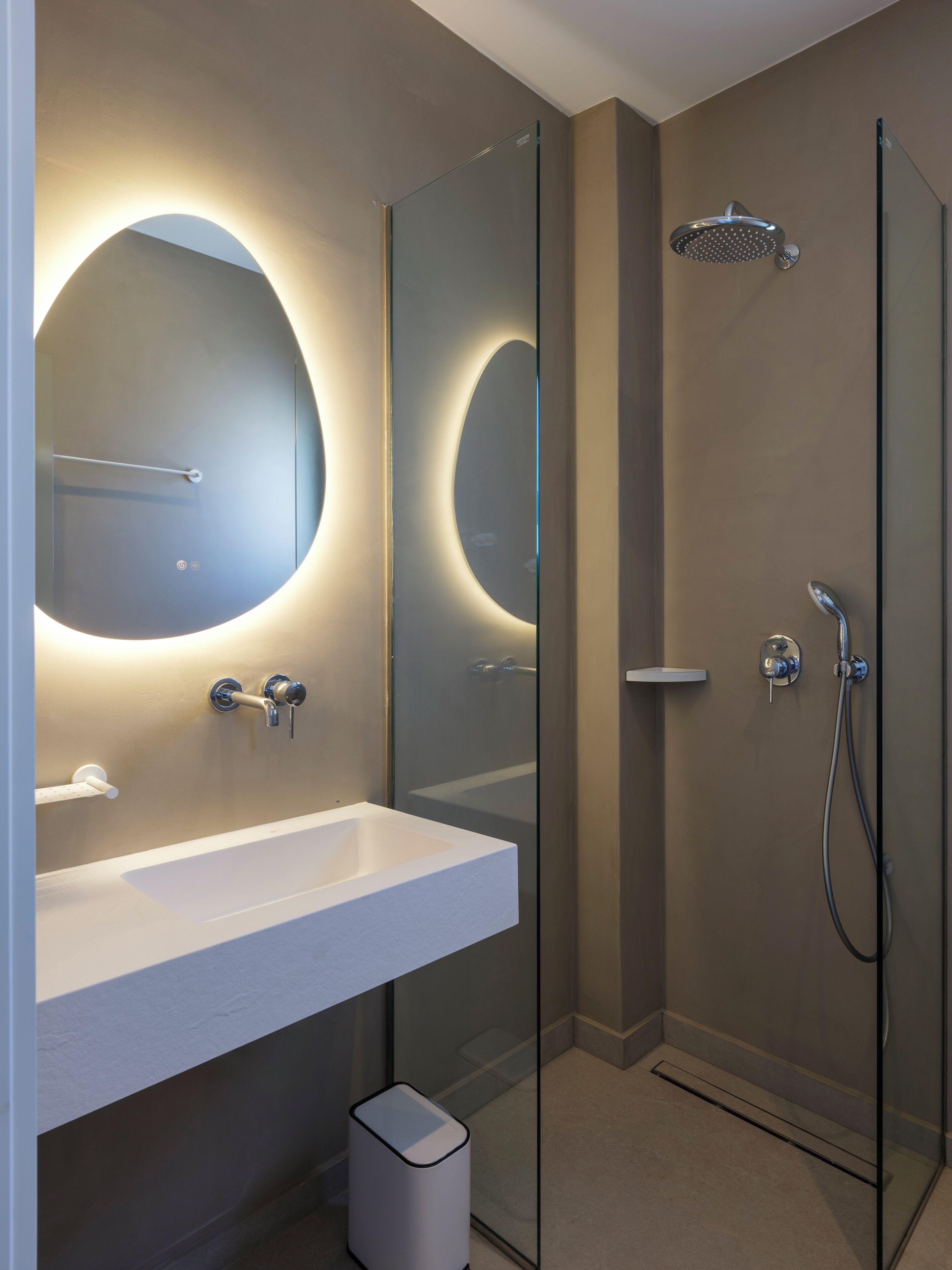 A modern bathroom with a glass shower enclosure, a white sink, and a large mirror with a circular light.