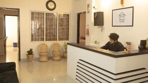 A reception desk with a clock, a window with blinds, and wicker chairs. - Malini Homes - Food & Lodging (Guwahati)