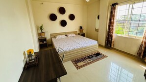 A bedroom with a bed, a desk, a ceiling fan, a window with curtains, and a view of greenery outside. - Malini Homes - Food & Lodging (Guwahati)