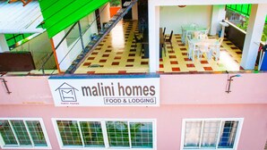 A building with a sign for 'Malini Homes Food & Lodging'. It features a checkered floor pattern, a staircase, and a dining area with tables and chairs. - Malini Homes - Food & Lodging (Guwahati)