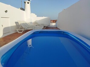 Une piscine sur le toit, avec des murs blancs, une cheminée blanche et plusieurs chaises longues blanches.