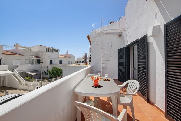Un balcon avec une table et des chaises, un mur blanc et une fenêtre avec des volets.